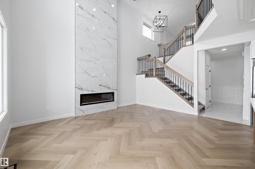 This expansive interior features a modern fireplace with a marble-style surround, herringbone pattern flooring, and a contemporary staircase with wood railings and black spindles - 236 Kinglet Boulevard Nw, Edmonton, AB - Indoor Photo Showing Other Room With Fireplace