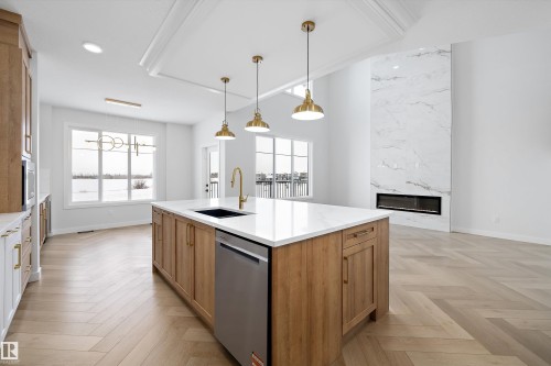 This open-concept living space features hardwood flooring laid in a herringbone pattern, a kitchen island with a white countertop and brass-toned fixtures, and a tall fireplace with a natural stone surround - 236 Kinglet Boulevard Nw, Edmonton, AB - Indoor Photo Showing Kitchen