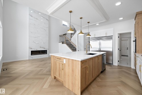 This expansive open-concept living area features a grand marble-style fireplace, light-toned chevron pattern flooring, and a kitchen island with a white countertop and brass-toned pendant lighting - 236 Kinglet Boulevard Nw, Edmonton, AB - Indoor Photo Showing Kitchen With Upgraded Kitchen