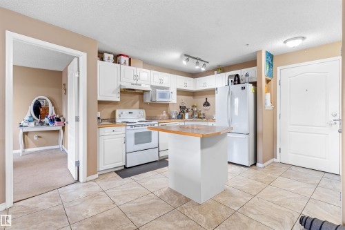 The property features a kitchen with white cabinetry, a central island with a wood countertop, and tile flooring - 224 3425 19 Street Nw, Edmonton, AB - Indoor Photo Showing Kitchen With Double Sink