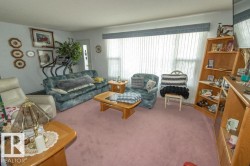 The living room features a broad window with blinds, carpeted flooring, and a light-colored wall - 