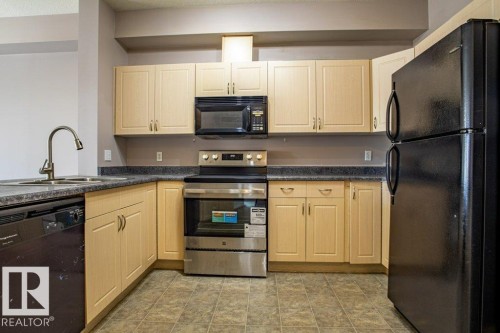 The kitchen features light wood cabinetry, dark countertops, a stainless steel range and microwave, and a black refrigerator and dishwasher - 326 4304 139 Ave, Edmonton, AB - Indoor Photo Showing Kitchen With Stainless Steel Kitchen With Double Sink