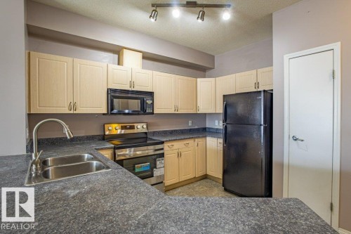 The kitchen features a double basin stainless steel sink, light wood cabinetry, dark countertops, and a black refrigerator - 326 4304 139 Ave, Edmonton, AB - Indoor Photo Showing Kitchen With Double Sink