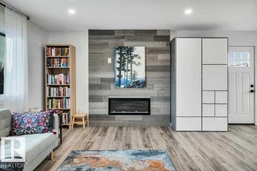 This living area features light wood-style flooring, a fireplace with a gray wood-paneled accent wall, and a white entry door with frosted glass detailing - 6719 94B Avenue, Edmonton, AB - Indoor Photo Showing Living Room With Fireplace