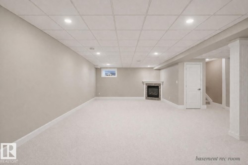 A spacious recreation room featuring neutral-toned carpeting, recessed lighting, and a corner fireplace with a stone surround - 6712 18 Ave Sw Sw, Edmonton, AB - Indoor