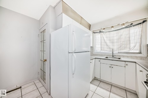 The kitchen features a white refrigerator, white cabinetry, a sink with a faucet, and a window with a curtain - 12411 126 Street Nw, Edmonton, AB - Indoor