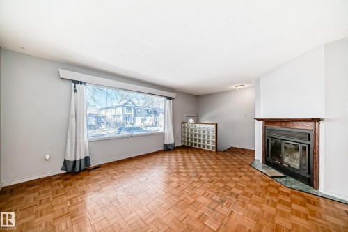 Living area featuring parquet wood flooring, a wide window with curtains, and a fireplace with a wood mantle and a dark stone hearth - 12411 126 Street Nw, Edmonton, AB - Indoor Photo Showing Living Room With Fireplace
