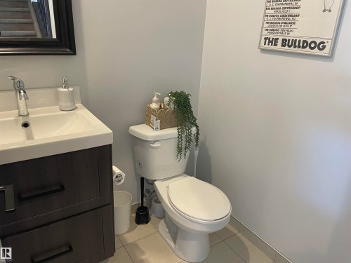 The bathroom features a modern vanity with a white countertop and dark cabinetry, a white toilet, and light-colored tiled flooring - 15220 105 Avenue, Edmonton, AB - Indoor Photo Showing Bathroom