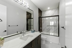 Contemporary bathroom featuring a vanity with a white countertop, a large mirror with integrated lighting, and a bathtub with a sliding glass door and tiled surround - 