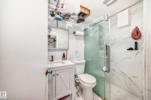 The bathroom features a vanity with a white countertop and cabinet, a toilet, and a glass-enclosed shower with white marble-patterned walls - 16305 107A Ave, Edmonton, AB - Indoor Photo Showing Bathroom