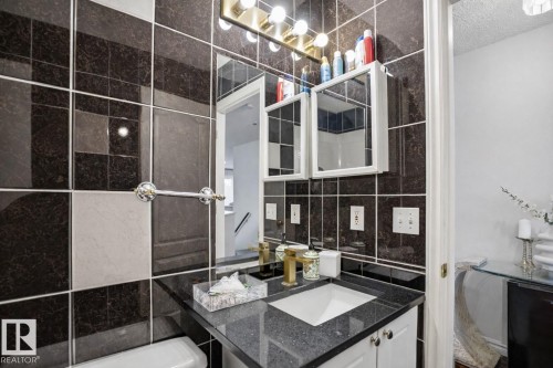 Bathroom featuring dark tiled walls with light accent tiles, a vanity with a dark speckled countertop, and a gold-toned faucet - 4619 202 Street, Edmonton, AB - Indoor Photo Showing Bathroom