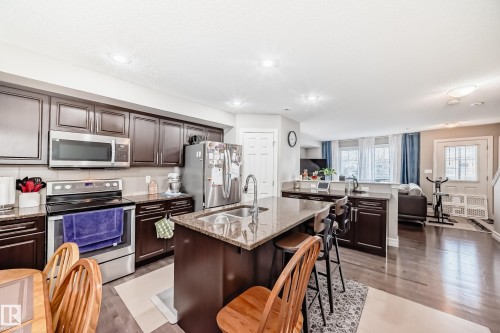 The kitchen features dark wood cabinetry, stainless steel appliances, granite countertops, and a central island with a sink and seating - 138 655 Tamarack Road Nw, Edmonton, AB - Indoor Photo Showing Kitchen With Double Sink With Upgraded Kitchen