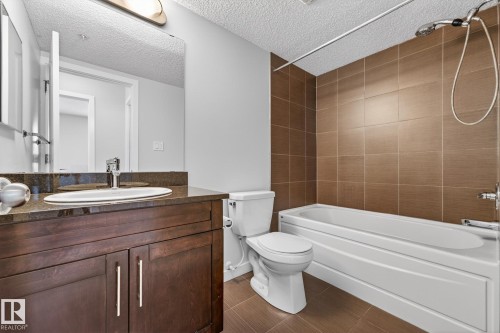 Bathroom featuring a dark wood vanity with a white oval sink and chrome faucet, a large mirror, a toilet, and a bathtub with brown tile surround - 301 1070 Mcconachie Boulevard, Edmonton, AB - Indoor Photo Showing Bathroom
