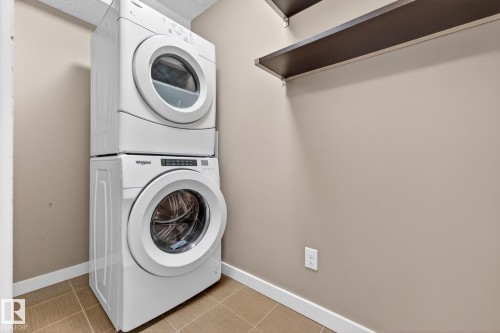 Laundry area featuring a stacked washer and dryer, tile flooring, and a wall-mounted shelf - 301 1070 Mcconachie Boulevard, Edmonton, AB - Indoor Photo Showing Laundry Room