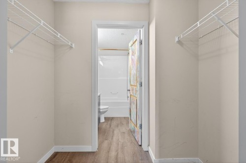 This area features wire shelving and wood-look flooring, with a view into a bathroom with a white toilet and tub - 109 340 Windermere Road, Edmonton, AB 