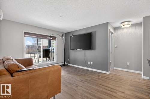 This living area features light wood-look flooring, light grey walls, and a sliding glass door providing access to a patio - 109 340 Windermere Road, Edmonton, AB 