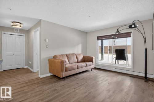Living area featuring wood-look flooring, light-colored walls, and a large window with horizontal blinds - 109 340 Windermere Road, Edmonton, AB 