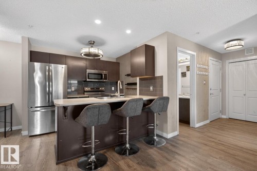 The kitchen features stainless steel appliances, dark wood cabinetry, and a light-colored countertop with an integrated sink and breakfast bar - 109 340 Windermere Road, Edmonton, AB 