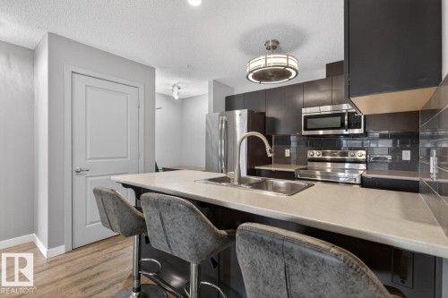 Modern kitchen featuring dark cabinetry, stainless steel appliances, a subway tile backsplash, and a large island with a sink and seating - 109 340 Windermere Road, Edmonton, AB 