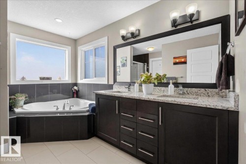 Bathroom featuring a corner bathtub with dark tile surround, dual windows, a long vanity with dual sinks, and a large mirror - 606 Reynalds Way, Leduc, AB - Indoor Photo Showing Bathroom