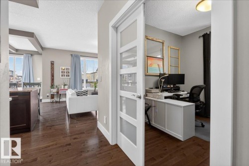 This property features an office space with a white desk and a door with frosted glass panels, alongside a living area with rich hardwood flooring - 606 Reynalds Way, Leduc, AB - Indoor