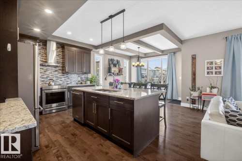 The kitchen features a large island with a granite countertop, dark wood cabinetry, stainless steel appliances, and wood flooring - 606 Reynalds Way, Leduc, AB - Indoor Photo Showing Kitchen With Upgraded Kitchen
