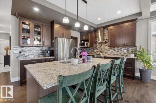 The kitchen features a large island with a light-colored countertop, dark wood cabinetry, a tiled backsplash, and stainless steel appliances - 606 Reynalds Way, Leduc, AB - Indoor