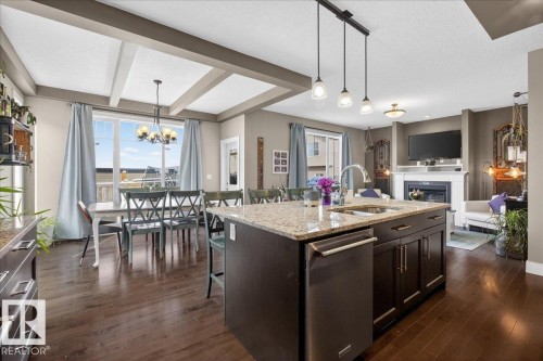 This open-concept living area features rich hardwood flooring, a kitchen island with granite countertops, and a dining area with a coffered ceiling - 606 Reynalds Way, Leduc, AB - Indoor Photo Showing Other Room