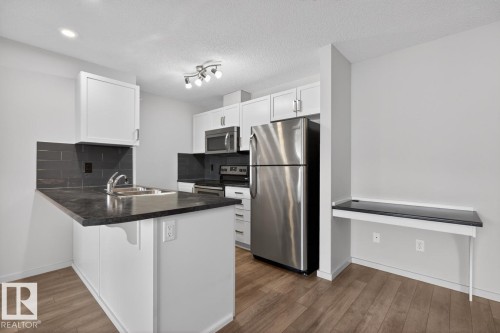 The kitchen features white cabinetry, stainless steel appliances, a dark countertop, and a built-in desk area - 426 390 Windermere Road, Edmonton, AB - Indoor Photo Showing Kitchen With Stainless Steel Kitchen With Double Sink