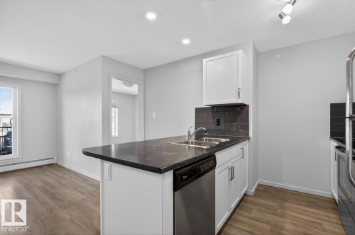 The kitchen features a functional island with a double stainless steel sink, a dishwasher, and a dark countertop - 426 390 Windermere Road, Edmonton, AB - Indoor Photo Showing Kitchen With Double Sink