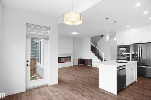 Open concept living area with hardwood style flooring, a modern fireplace, and a kitchen featuring a white island with a sink and pendant lighting - Edmonton, AB - Indoor Photo Showing Kitchen With Fireplace