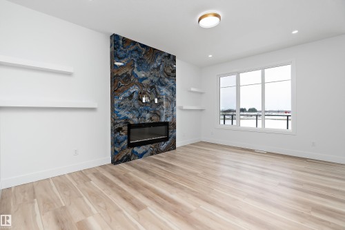 Living area featuring light wood flooring, a fireplace with a striking blue and brown patterned surround, and large windows providing views of the outdoors - 219 Crystal Creek Dr, Leduc, AB - Indoor Photo Showing Living Room With Fireplace