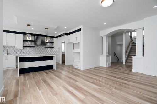 Open-concept living space with light wood-style flooring, a modern kitchen featuring a white and dark blue island, white cabinetry, and a geometric tile backsplash, and a staircase with light wood banister - 219 Crystal Creek Dr, Leduc, AB - Indoor Photo Showing Kitchen
