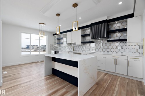 The kitchen features a large island with a quartz countertop, white cabinetry with gold-toned hardware, a distinctive black and white patterned backsplash, and light wood flooring - 219 Crystal Creek Dr, Leduc, AB - Indoor Photo Showing Kitchen With Upgraded Kitchen