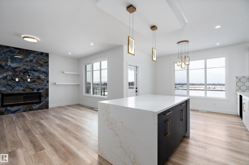 This open-concept area features light wood-style flooring, a kitchen island with a white countertop and dark cabinetry, and multiple large windows - 219 Crystal Creek Dr, Leduc, AB - Indoor With Fireplace