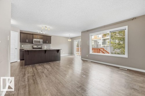 This open concept interior features wood-look flooring, a kitchen island with dark cabinetry, and a large window providing views of the outdoor deck and surrounding properties - 7873 170A Avenue, Edmonton, AB - Indoor
