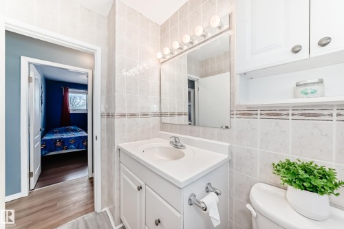 The bathroom features a vanity with a white countertop and an integrated sink, a wall-mounted mirror with overhead lighting, and tiled walls with a decorative accent band - 7512 136 Avenue, Edmonton, AB - Indoor Photo Showing Bathroom