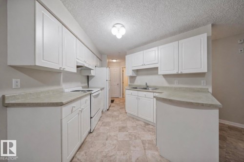 The kitchen features white cabinetry, light countertops, and a full suite of white appliances including a refrigerator and range - 103 12110 119 Ave, Edmonton, AB - Indoor Photo Showing Kitchen With Double Sink