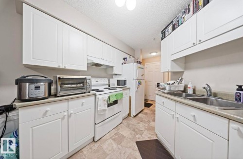 The kitchen features white cabinetry, a double basin sink, and a refrigerator - 103 12110 119 Ave, Edmonton, AB - Indoor Photo Showing Kitchen With Double Sink
