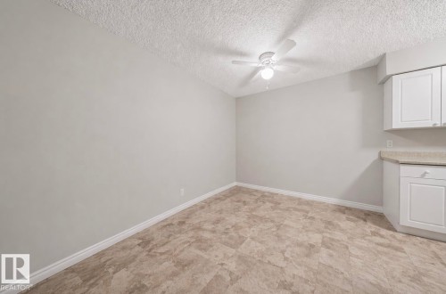 This room features neutral-toned walls, tile-patterned flooring, a ceiling fan, and white cabinetry with a light-colored countertop - 103 12110 119 Ave, Edmonton, AB - Indoor
