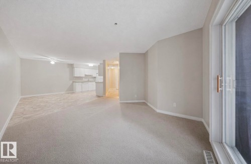 Spacious open-concept living area featuring light-colored carpeting, neutral painted walls, and a sliding glass door - 103 12110 119 Ave, Edmonton, AB - Indoor Photo Showing Other Room
