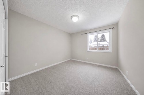 This room features light gray carpeting, neutral-colored walls, and a window with a view of a residential area with trees - 103 12110 119 Ave, Edmonton, AB - Indoor Photo Showing Other Room