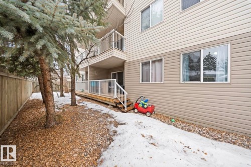 Exterior view of the property featuring a balcony with white railings, a ground-level patio, and a fenced yard with mature trees - 103 12110 119 Ave, Edmonton, AB - Outdoor With Balcony With Exterior