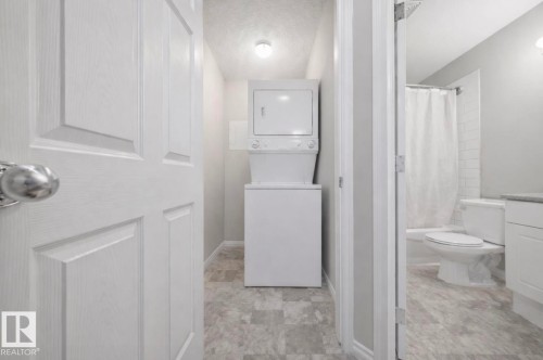 Compact laundry area with a stackable washer and dryer, adjacent to a bathroom featuring a toilet and a shower with a white curtain - 103 12110 119 Ave, Edmonton, AB - Indoor