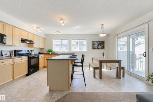 The kitchen features light wood cabinetry, black appliances, a central island with seating, and tiled flooring - 67 12050 17 Avenue, Edmonton, AB - Indoor Photo Showing Kitchen