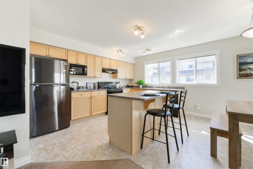 The kitchen features light-toned cabinetry, a central island with a breakfast bar, and two windows providing natural illumination - 67 12050 17 Avenue, Edmonton, AB - Indoor Photo Showing Kitchen