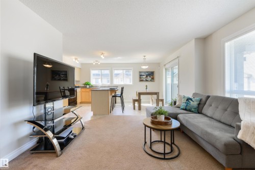 This bright open-concept living space features carpeted flooring, white walls, and a large window with blinds - 67 12050 17 Avenue, Edmonton, AB - Indoor Photo Showing Living Room