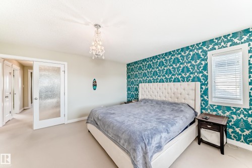 This room features a decorative accent wall with a blue and white patterned wallpaper, a light-colored carpet, and a window with blinds - 3433 Goodridge Link Link, Edmonton, AB - Indoor Photo Showing Bedroom