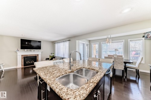 This open-concept living space features hardwood floors, a kitchen island with a double sink and granite countertop, a fireplace with a mantel, and large windows in the dining area - 3433 Goodridge Link Link, Edmonton, AB - Indoor Photo Showing Kitchen With Fireplace With Double Sink