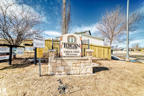 Entrance to Tuscan Village, featuring a stone and iron sign with the community name and address, surrounded by dry grass and bare trees - 44 13825 155 Avenue, Edmonton, AB - Outdoor